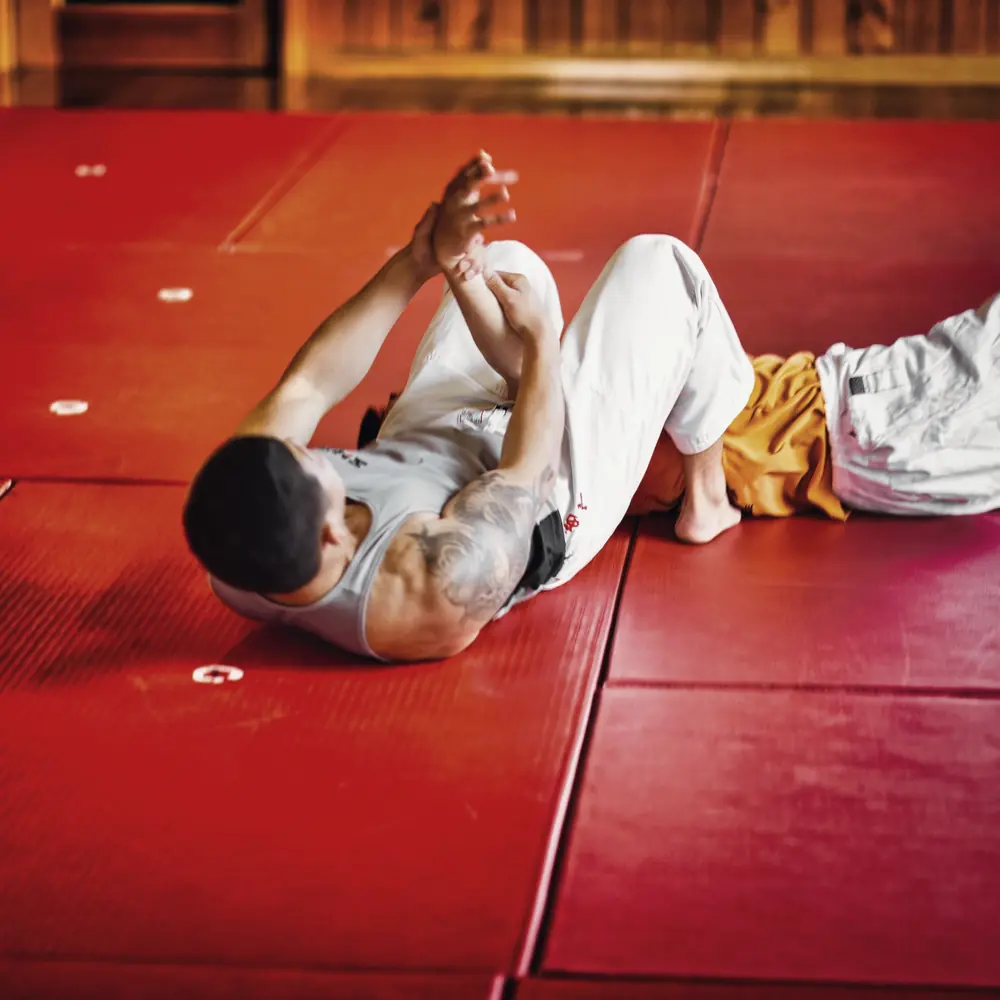A club member on his back on the red mats, hands clasped, mid-way through a grappling hold during practice.