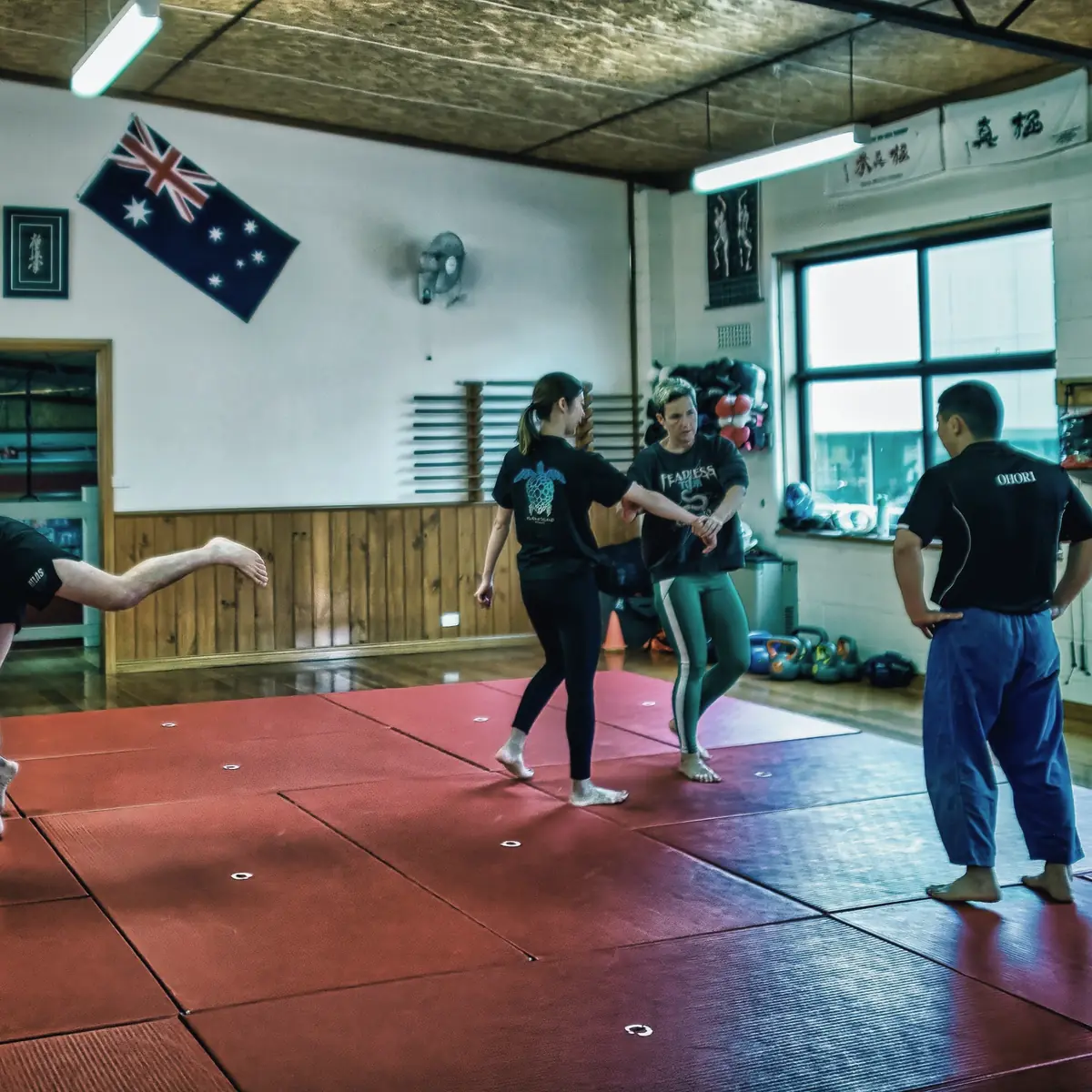 Two club members practise a standing grip drill on the red mats while a coach looks on, inside the Percy Street dojo.
